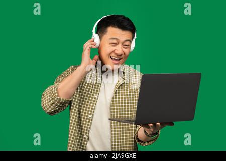 Felice uomo asiatico maturo in cuffie wireless utilizzando un PC portatile per il lavoro online o la comunicazione, sfondo verde Foto Stock