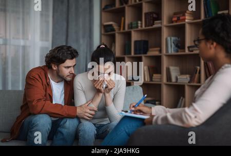 Il giovane europeo disperato calma piangendo la signora afroamericana su consultazione con lo psicologo in clinica Foto Stock