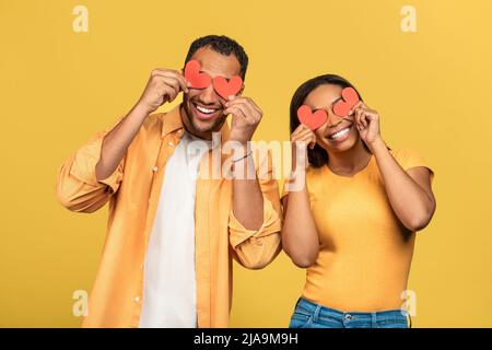 Accecato dal concetto di amore. Affettuosa coppia afroamericana che copre gli occhi con cuori di carta rossa su sfondo giallo Foto Stock