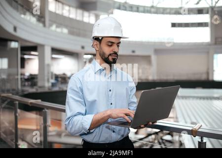 Tecnico di tipo arabo pensivo occupato millennial in casco di sicurezza con barba lavora con computer controlli fabbrica Foto Stock