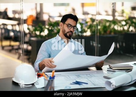 Serio bel giovane ingegnere musulmano in occhiali con barba con disegni di progetto sul posto di lavoro con elmetto Foto Stock