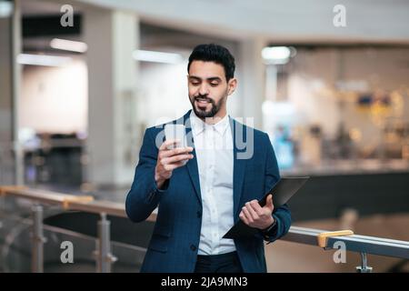 Allegro e bello uomo d'affari arabo millenario con la barba in tuta con i look tablet dello smartphone in ufficio Foto Stock