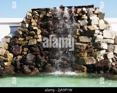 Una cascata scorre da un muro di pietra, cadendo in una piscina turchese. Area decorativa estiva per passeggiate Foto Stock
