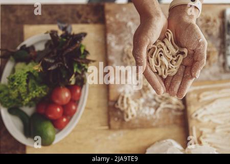 Tagliatelle fatte in casa nelle mani delle donne su un tavolo da cucina con farina e verdure su un piatto Foto Stock