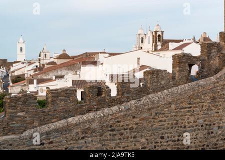 Uomo in camicia bianca che cammina sui bastioni del castello di Monsaraz, case bianche sullo sfondo. Alentejo, Portogallo. Foto Stock