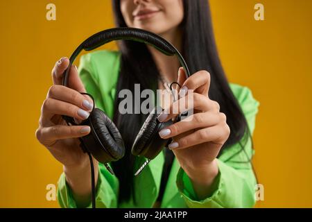 Vista rifilata di una donna sorridente con mani pulite che tengono in mano gli auricolari neri in studio. Vista ravvicinata delle cuffie che reggono una donna felice in un blazer verde, isolato su sfondo arancione. Concetto di musica. Foto Stock