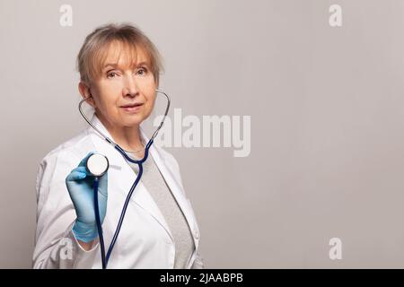 Medico donna matura in uniforme medica in piedi e stetoscopio di tenuta su sfondo grigio studio. Infermiere medico femminile o ritratto medico. Correggi Foto Stock