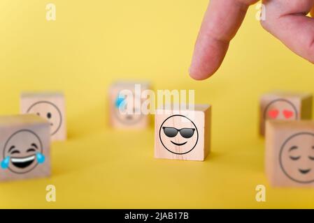 Il dito punta a un'emoji sorridente con gli occhiali su sfondo giallo. Il concetto della giornata mondiale delle emoticon. Foto Stock