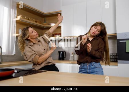 Donna e ragazza ballano, cantando con spatole in legno come microfono in cucina e in cucina. Mamma e figlia si divertono Foto Stock