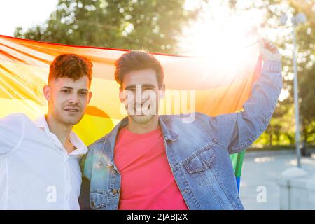 Giovane coppia gay che tiene una bandiera arcobaleno LGBT mentre si alza insieme all'aperto. LGBT, concetto di relazione e parità di diritti. Foto Stock