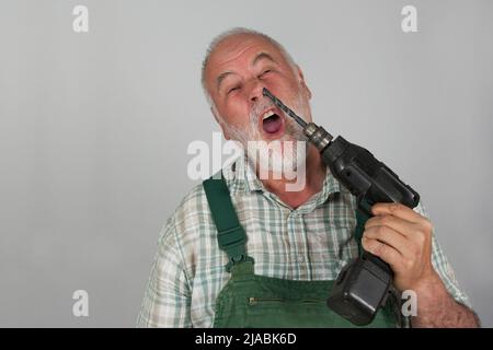 Presa letteralmente nel naso che raccoglie. Un vecchio artigiano si afferra al naso con la sua macchina da perforazione, di fronte ad un muro bianco. Foto Stock