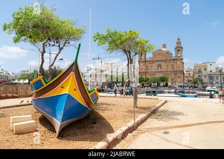 Msida, Malta - Maggio 29th 2022: Il peschereccio maltese chiamò Luzzu su terra asciutta con la chiesa parrocchiale di San Giuseppe sullo sfondo. Foto Stock