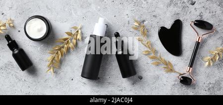 Piatto laico nero obsidian viso rullo, gua sha, siero, oli essenziali e rami d'oro su tavola di pietra. Set cosmetici per la cura della pelle di lusso. Foto Stock