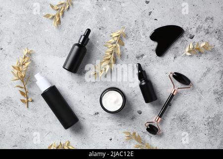 Flat Lay lusso viso cura della pelle cosmetici. Vista dall'alto rullo per massaggio facciale, gua sha, vaso di idratante, siero, oli essenziali e rami d'oro su co Foto Stock