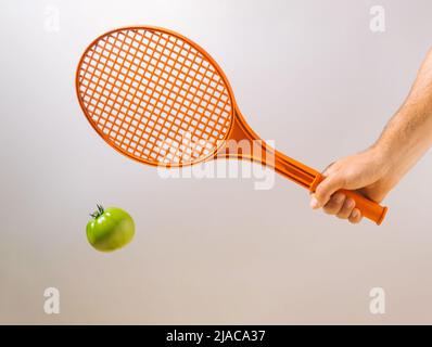 Mano che tiene una racchetta da tennis che colpisce un pomodoro verde non maturo su sfondo grigio. Concetto sportivo minimo. Foto Stock
