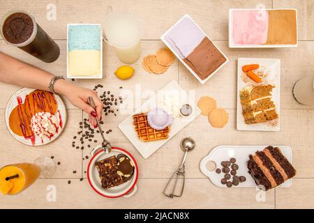 Vista dall'alto di un set di piatti da dessert, gelato, torta al cioccolato, mano con cucchiaio, chicchi di caffè, pancake con panna, gelato assortito Foto Stock