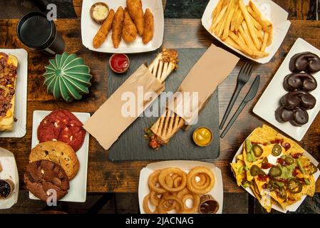 Set di piatti fast food con patatine fritte, patatine dolci, biscotti al cioccolato, nachos con guacamole, anelli di cipolla, panini e salse varie, Foto Stock