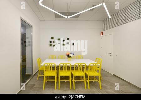 Grande tavolo da pranzo rettangolare di legno bianco circondato da sedie gialle con luci fluorescenti sul soffitto e accesso alla terrazza Foto Stock