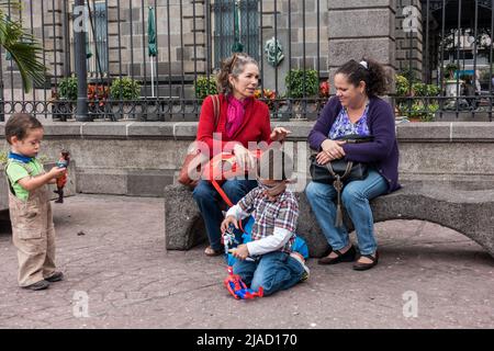 Due donne che parlano seduti su una panca del parco mentre due bambini giocano con i loro giocattoli a San José, Costa Rica. Foto Stock