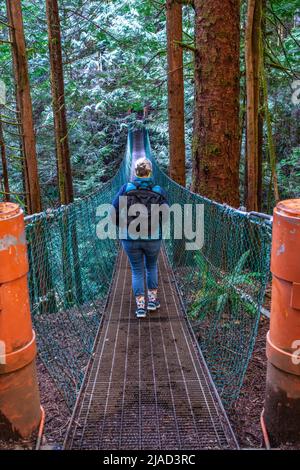 Vista posteriore di una donna che attraversa un ponte sospeso, Juan de Fuca Marine Trail, Juan de Fuca Provincial Park, Vancouver Island, British Columbia, Canada Foto Stock