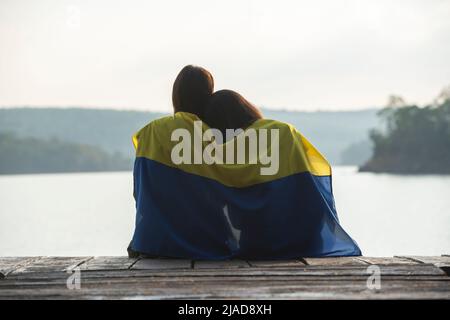 Vista posteriore di una coppia seduta su un molo avvolto in una bandiera Ucraina, Thailandia Foto Stock