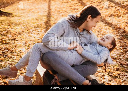 Giovane madre che conforta la figlia mentre la tiene in grembo seduto su una panca nel parco. Buona famiglia insieme. Cura della famiglia. Foto Stock