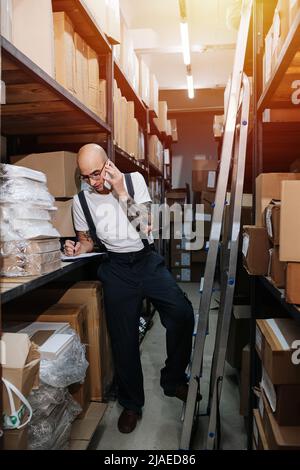 Lavoratore di magazzino maschile sovraccarichi di lavoro che parla al telefono, scrivendo su una clipboard. Si trova tra file alte di scaffali con scatole. Foto Stock