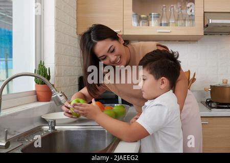 Madre sorridente che mostra a figlio piccolo come lavare la frutta sotto l'acqua del rubinetto prima di mangiare Foto Stock