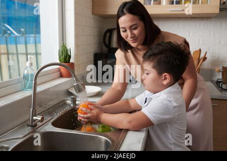 Bambino che sciacqua la frutta sotto l'acqua del rubinetto sotto il controllo della madre Foto Stock