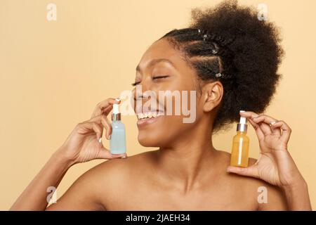 Bella donna afroamericana che tiene bottiglie di acido ialuronico per la cura della pelle facciale e siero di vitamina C. Donna nera che tiene vasi di vetro isolato o Foto Stock