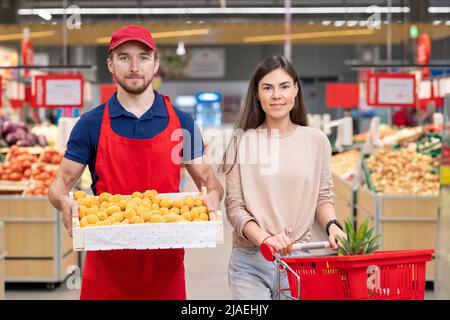 Colpo medio orizzontale di lavoratore maschio che tiene scatola di albicocche stare in piedi con una giovane cliente femminile nella corsia del supermercato Foto Stock