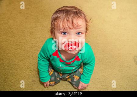 allegro bambino sorridente dai capelli rossi siede sul pavimento con un succhietto in bocca Foto Stock