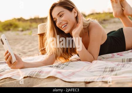 Giovane donna bionda sorridente e con cellulare mentre si trova sulla spiaggia Foto Stock