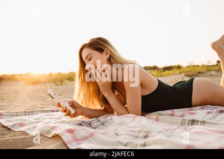 Giovane donna bionda sorridente e con cellulare mentre si trova sulla spiaggia Foto Stock