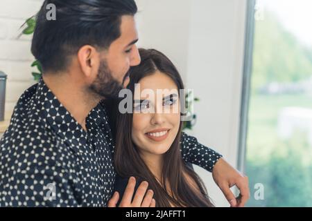 Attraente uomo bearded abbraccia una bella donna attraente e guarda la finestra, mentre la donna guarda la fotocamera e sorridendo ampiamente. Foto di alta qualità Foto Stock