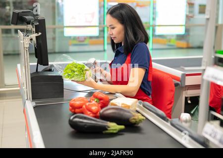 Attraente cassiere asiatico con grembiule rosso che vende verdure fresche al cliente Foto Stock