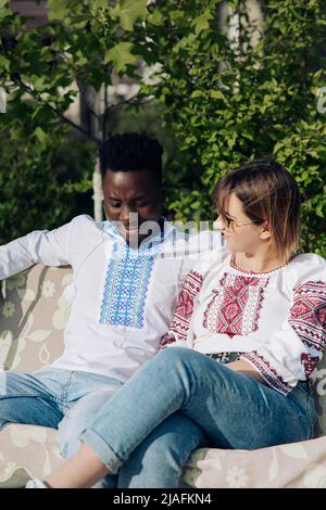 Coppia interrazziale si siede su panca in giardino vestito di magliette tradizionali ucraine ricamate etniche. Concetto di relazioni d'amore e di unità tra Foto Stock