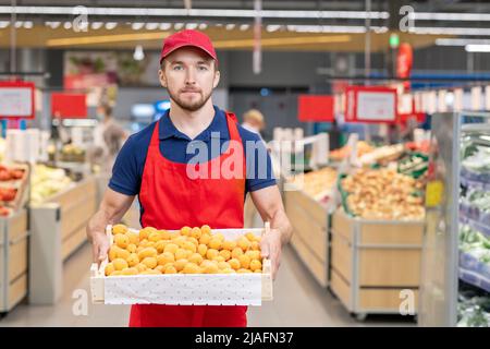 Colpo medio orizzontale di lavoratore maschio che tiene scatola di albicocche stare in piedi con una giovane cliente femminile nella corsia del supermercato Foto Stock