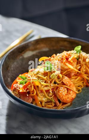 Mescolare le tagliatelle con verdure, gamberetti e frutti di mare in una ciotola nera su un tavolo di marmo. Foto Stock