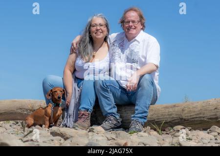 Coppia escursionistica matura seduta su un ceppo facendo una pausa con il loro dachshund marrone, riserva naturale Maasvallei, cielo blu sullo sfondo, giorno di sole in S Foto Stock