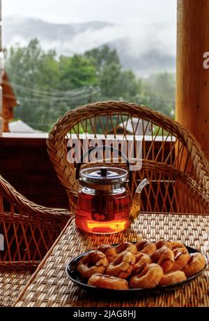 Tè di frutta calda in una teiera di vetro e biscotti si trova sul bordo di un tavolo di rattan. Foto Stock