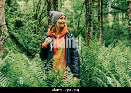 Giovane donna attiva escursionista in giacca e un cappello con uno zaino in un thicket di felci nella foresta Foto Stock