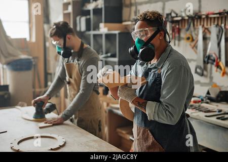 Ritratto laterale di due falegnami in attrezzatura protettiva che lavorano con legno mentre costruisce mobili artigianali in officina Foto Stock