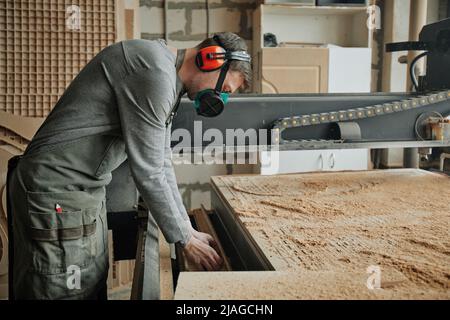 Ritratto della vista laterale del lavoratore maschile che indossa un equipaggiamento protettivo completo durante il funzionamento delle macchine nella produzione di legno Foto Stock