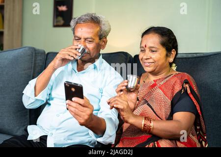 sorridente coppia anziana che usa il telefono cellulare mentre ha il tè a casa sul divano - concetto di relax Foto Stock