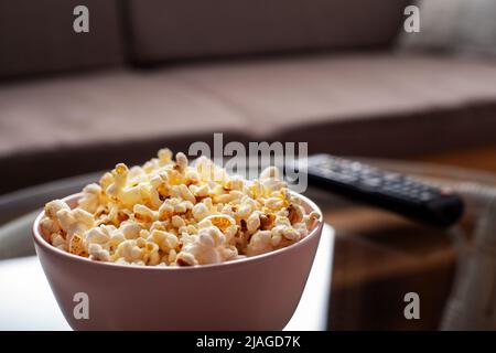 Ciotola con popcorn e telecomando tv su tavolo in vetro vicino al divano Foto Stock
