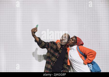 Uomo felice che prende selfie con la figlia che sbuffa le labbra mentre si alza contro il muro durante la giornata di sole Foto Stock