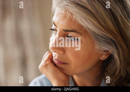 Primo piano Shot di una donna matura pensierosa che guarda fuori dalla finestra a casa Foto Stock