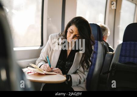 Donna d'affari che scrive nel diario mentre si siede dalla finestra del treno Foto Stock