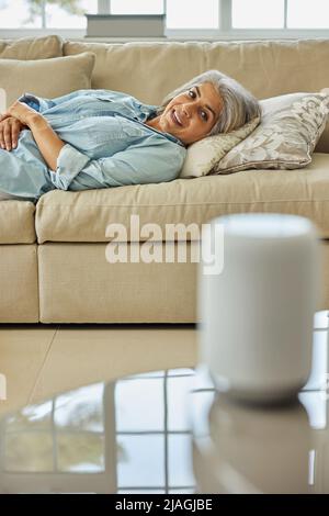 Donna matura sdraiata sul divano che pone una domanda all'assistente digitale o all'altoparlante intelligente Foto Stock